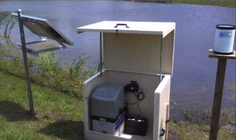Portable water sampler housed in a weatherproof outdoor enclosure beside a pond, connected to a solar panel and a nearby rain gauge.