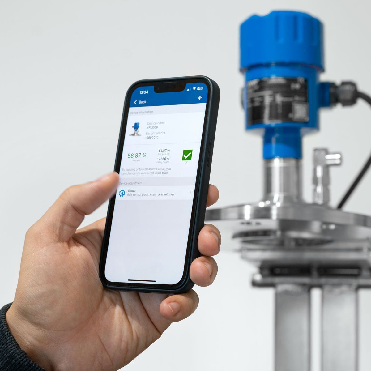 Hand holding smartphone displaying radar level transmitter data through a Bluetooth app, with the sensor visible in the background.